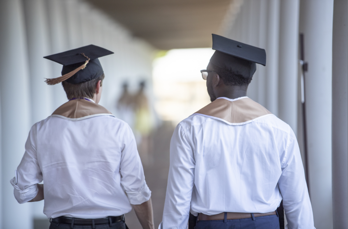 Two graduating students walking down a pillared walkway wearing graduation caps.